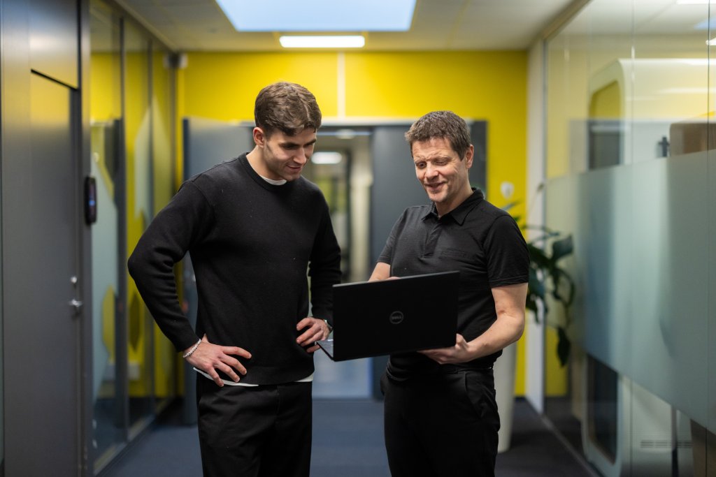 To personer står i korridoren, en av dem viser noe på laptopen og den andre ser.